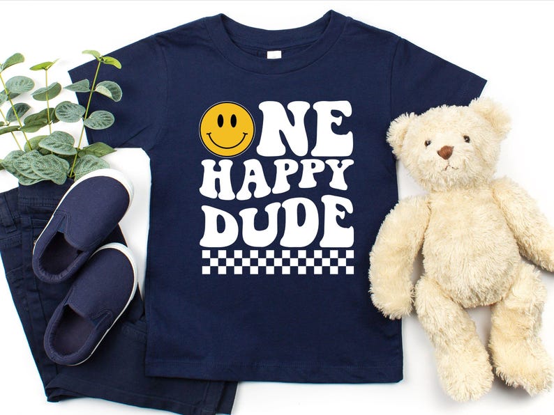 May include: Navy blue t-shirt with the text "ONE HAPPY DUDE" in white, featuring a yellow smiley face. The shirt is paired with navy blue pants and shoes, and a teddy bear. A checkered pattern is at the bottom of the shirt.