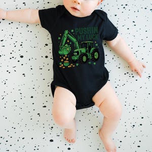 May include: Black baby bodysuit with a green tractor graphic, shamrocks, and gold coins. The text "Pushin' My Luck" is printed above the tractor. The bodysuit is designed for St. Patrick's Day.