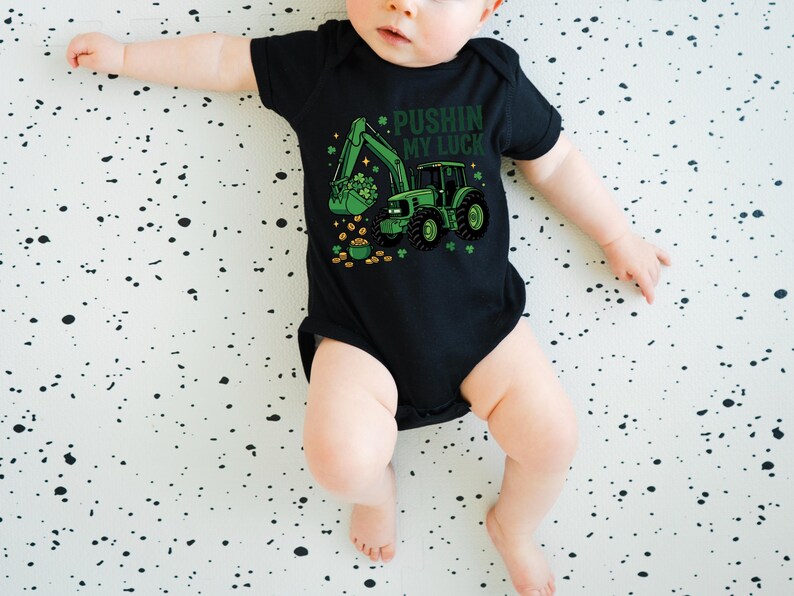 May include: Black baby bodysuit with a green tractor graphic, shamrocks, and gold coins. The text "Pushin' My Luck" is printed above the tractor. The bodysuit is designed for St. Patrick's Day.