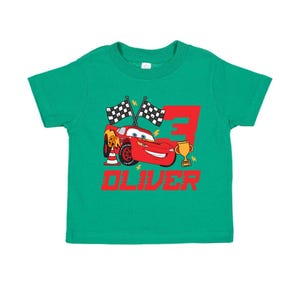 May include: A green toddler t-shirt featuring a red cartoon race car, a gold trophy, and the number 3. The name "OLIVER" is printed in red below the car. Two checkered flags are also included in the design.