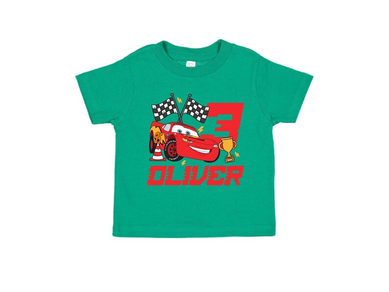 May include: A green toddler t-shirt featuring a red cartoon race car, a gold trophy, and the number 3. The name "OLIVER" is printed in red below the car. Two checkered flags are also included in the design.