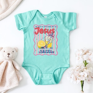 May include: A light turquoise baby bodysuit with a graphic of a glass of lemonade, a lemon slice, and the text "Jesus He Be Makin' Lemonade". A teddy bear and a white flower bouquet are next to the bodysuit.