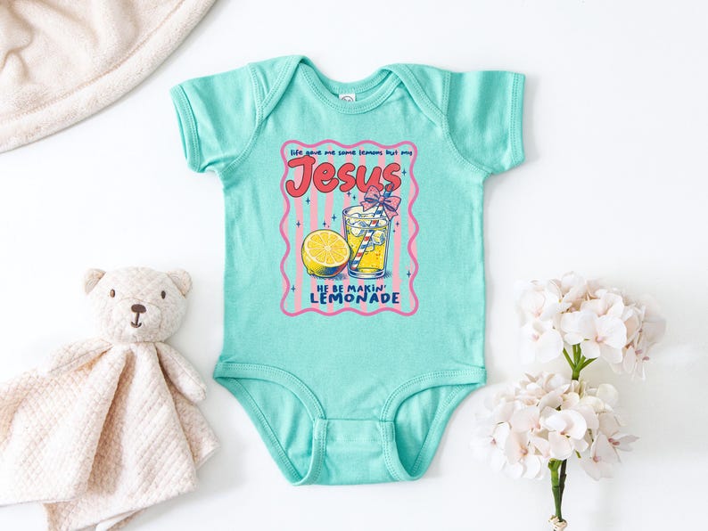 May include: A light turquoise baby bodysuit with a graphic of a glass of lemonade, a lemon slice, and the text "Jesus He Be Makin' Lemonade". A teddy bear and a white flower bouquet are next to the bodysuit.