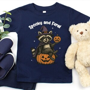 Peut inclure: T-shirt bleu marine avec un motif d'Halloween. Le motif repr&eacute;sente un raton laveur portant un chapeau de sorci&egrave;re et une cape, assis sur une citrouille d'Halloween et tenant une citrouille. Le texte "Spooky and Feral" est au-dessus du raton laveur. Comprend un pantalon et des chaussures bleu marine.