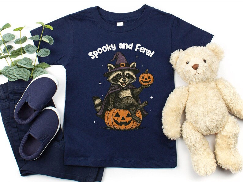 Peut inclure: T-shirt bleu marine avec un motif d'Halloween. Le motif repr&eacute;sente un raton laveur portant un chapeau de sorci&egrave;re et une cape, assis sur une citrouille d'Halloween et tenant une citrouille. Le texte "Spooky and Feral" est au-dessus du raton laveur. Comprend un pantalon et des chaussures bleu marine.