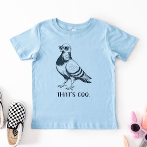 May include: A light blue t-shirt featuring a black and white illustration of a pigeon wearing sunglasses. The text "THAT'S COO." is below the bird. A pair of black and white checkered slip-on shoes are to the left.
