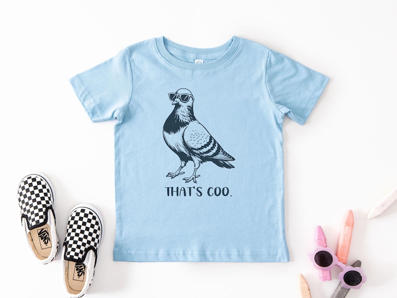 May include: A light blue t-shirt featuring a black and white illustration of a pigeon wearing sunglasses. The text "THAT'S COO." is below the bird. A pair of black and white checkered slip-on shoes are to the left.