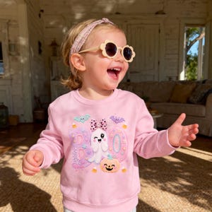 Puede incluir: Una sudadera de manga larga rosa claro con un estampado gr&aacute;fico que incluye la palabra BOO, un fantasma gui&ntilde;ando un ojo con un lazo de lunares, una calabaza, murci&eacute;lagos y caramelos. Se combina con gafas de sol color crema en forma de flor.