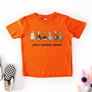 May include: Orange short-sleeved t-shirt with a graphic of six cartoon geese dressed in Halloween costumes. The text "just a spooky goose" is below the geese. Black and white checkered slip-on shoes are on the left.