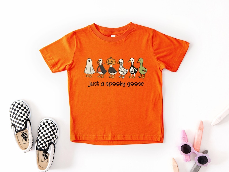 May include: Orange short-sleeved t-shirt with a graphic of six cartoon geese dressed in Halloween costumes. The text "just a spooky goose" is below the geese. Black and white checkered slip-on shoes are on the left.