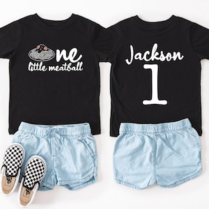 May include: Two black t-shirts with white text and light blue denim shorts. One shirt says "one little meatball" with a spaghetti and meatball graphic. The other shirt says "Jackson 1". Checkered slip-on shoes are also included.