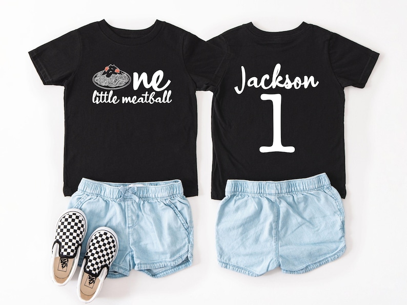 May include: Two black t-shirts with white text and light blue denim shorts. One shirt says "one little meatball" with a spaghetti and meatball graphic. The other shirt says "Jackson 1". Checkered slip-on shoes are also included.