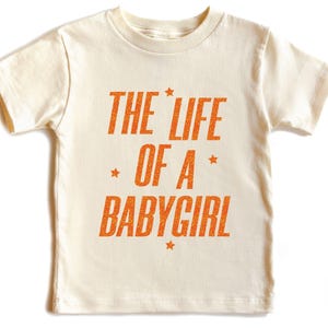 Pode incluir: T-shirt creme para crian&ccedil;a com o texto laranja "THE LIFE OF A BABYGIRL", adornado com estrelas. O texto est&aacute; em negrito. A t-shirt de manga curta tem gola redonda.