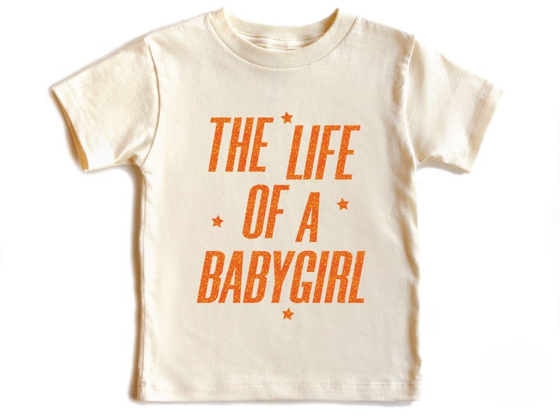 Pode incluir: T-shirt creme para crian&ccedil;a com o texto laranja "THE LIFE OF A BABYGIRL", adornado com estrelas. O texto est&aacute; em negrito. A t-shirt de manga curta tem gola redonda.