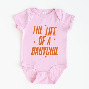 Pode incluir: Body de beb&eacute; rosa claro com o texto laranja "THE LIFE OF A BABYGIRL". O texto tem um aspeto desgastado com detalhes de estrelas. &Eacute; uma pe&ccedil;a de roupa de beb&eacute; de uma pe&ccedil;a.