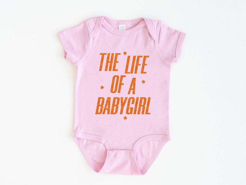 Pode incluir: Body de beb&eacute; rosa claro com o texto laranja "THE LIFE OF A BABYGIRL". O texto tem um aspeto desgastado com detalhes de estrelas. &Eacute; uma pe&ccedil;a de roupa de beb&eacute; de uma pe&ccedil;a.