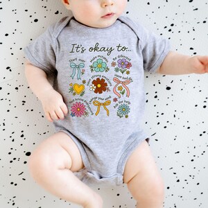 May include: A gray baby onesie with the text "It's okay to..." and colorful floral and ribbon graphics. The design includes phrases like "Start over," "Make mistakes," and "Have strong emotions."