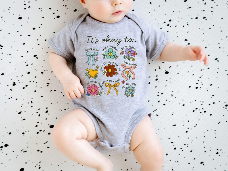 May include: A gray baby onesie with the text "It's okay to..." and colorful floral and ribbon graphics. The design includes phrases like "Start over," "Make mistakes," and "Have strong emotions."