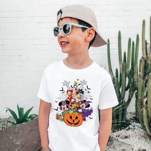 Op de afbeelding: Wit T-shirt met een Halloween-thema met stripfiguren, waaronder Mickey en Minnie Mouse, Donald Duck en Goofy, rond een uitgesneden pompoen. Het ontwerp bevat vleermuizen en spinnenwebben. Het kind draagt een pet en een zonnebril.