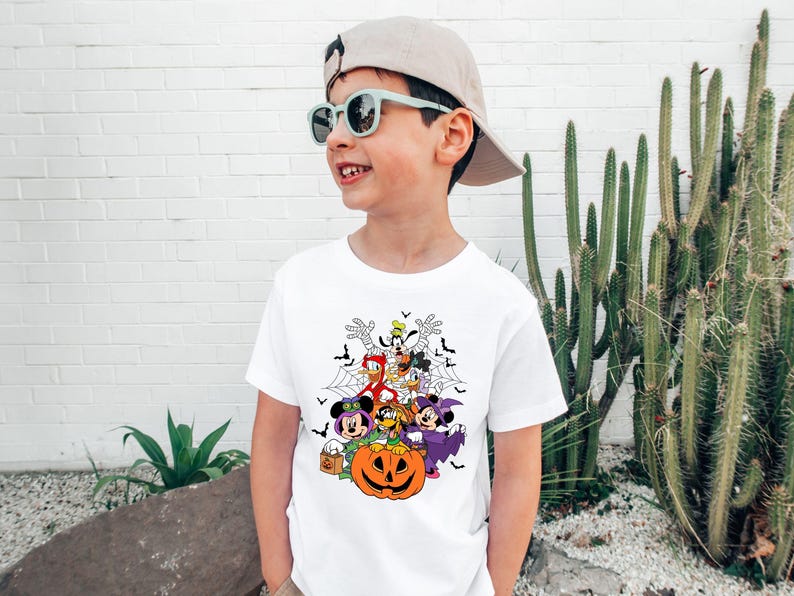 Op de afbeelding: Wit T-shirt met een Halloween-thema met stripfiguren, waaronder Mickey en Minnie Mouse, Donald Duck en Goofy, rond een uitgesneden pompoen. Het ontwerp bevat vleermuizen en spinnenwebben. Het kind draagt een pet en een zonnebril.