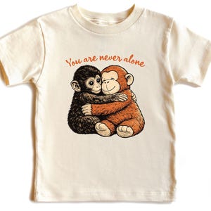 May include: Cream-colored t-shirt featuring a graphic of two hugging monkeys. One monkey is brown, the other is black. The text "You are never alone" is printed above the monkeys in orange.