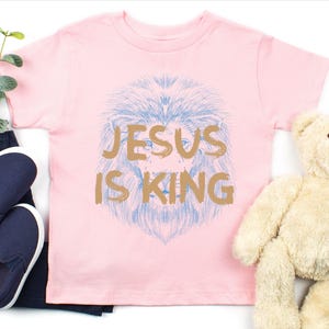 May include: A light pink t-shirt with the text "JESUS IS KING" in gold lettering. The shirt features a blue lion's head design. A pair of navy blue slip-on shoes and a teddy bear are also in the image.