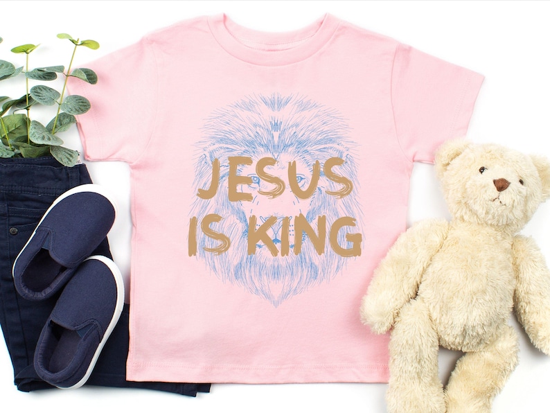 May include: A light pink t-shirt with the text "JESUS IS KING" in gold lettering. The shirt features a blue lion's head design. A pair of navy blue slip-on shoes and a teddy bear are also in the image.
