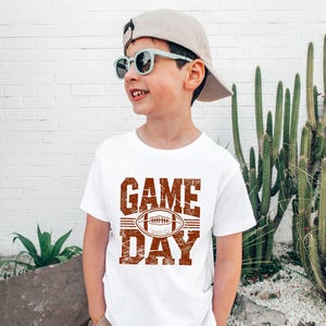 K&ouml;nnte beinhalten: Ein wei&szlig;es T-Shirt mit einem verwitterten braunen "GAME DAY"-Aufdruck und einer Football-Illustration. Das T-Shirt wird von einer Person mit Sonnenbrille und Baseballkappe getragen. Der Hintergrund umfasst eine wei&szlig;e Backsteinmauer und Kakteen.
