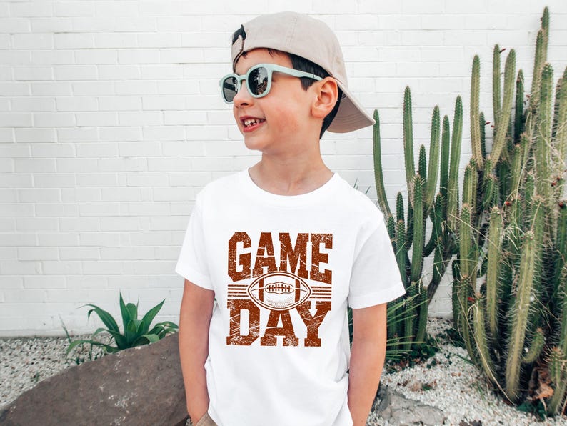 K&ouml;nnte beinhalten: Ein wei&szlig;es T-Shirt mit einem verwitterten braunen "GAME DAY"-Aufdruck und einer Football-Illustration. Das T-Shirt wird von einer Person mit Sonnenbrille und Baseballkappe getragen. Der Hintergrund umfasst eine wei&szlig;e Backsteinmauer und Kakteen.