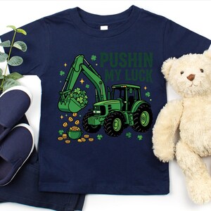 May include: Navy blue toddler t-shirt with a green tractor graphic, a bucket full of shamrocks, and the text "Pushin' My Luck". The design includes gold coins and shamrocks. The shirt is paired with navy blue pants and shoes, and a teddy bear.