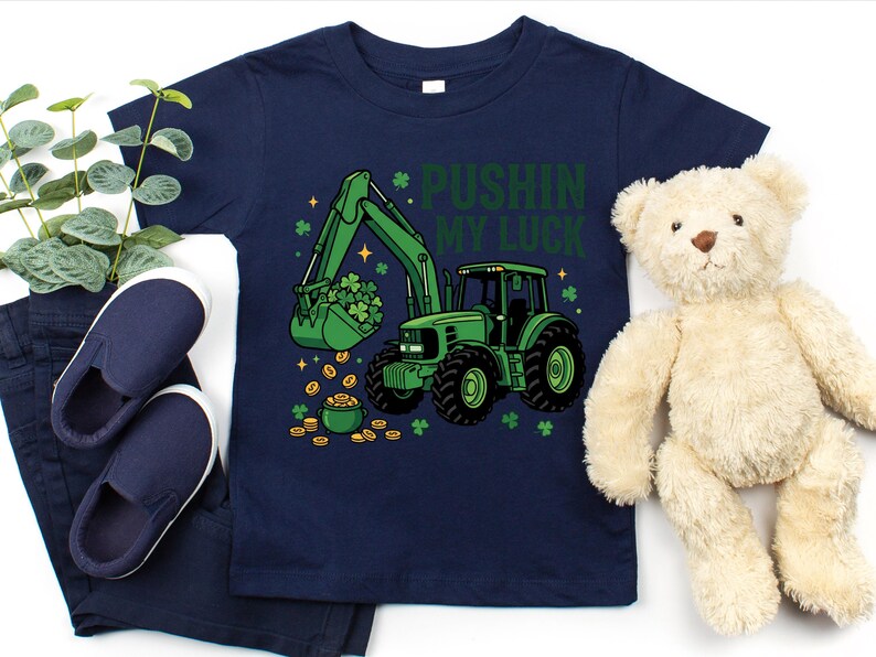 May include: Navy blue toddler t-shirt with a green tractor graphic, a bucket full of shamrocks, and the text "Pushin' My Luck". The design includes gold coins and shamrocks. The shirt is paired with navy blue pants and shoes, and a teddy bear.
