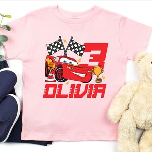 May include: A light pink t-shirt featuring a red cartoon race car with the number 3 and the name OLIVIA. The design includes checkered flags, a trophy, and lightning bolts. The shirt is paired with navy blue pants and shoes, and a teddy bear.
