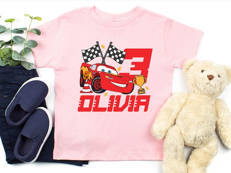 May include: A light pink t-shirt featuring a red cartoon race car with the number 3 and the name OLIVIA. The design includes checkered flags, a trophy, and lightning bolts. The shirt is paired with navy blue pants and shoes, and a teddy bear.