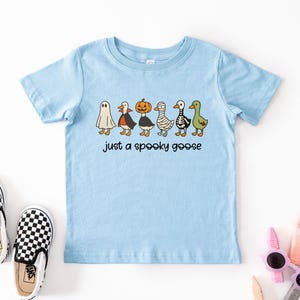 May include: Light blue short-sleeved t-shirt featuring a line of cartoon geese dressed in Halloween costumes, including a ghost, vampire, pumpkin, mummy, skeleton, and green goose. The text "just a spooky goose" is below the geese. Black and white checkered slip-on shoes are in the image.