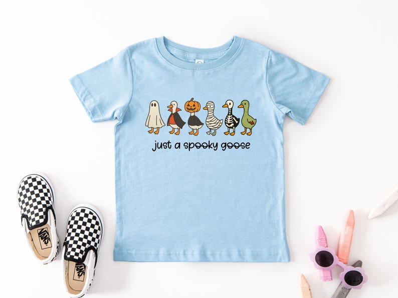 May include: Light blue short-sleeved t-shirt featuring a line of cartoon geese dressed in Halloween costumes, including a ghost, vampire, pumpkin, mummy, skeleton, and green goose. The text "just a spooky goose" is below the geese. Black and white checkered slip-on shoes are in the image.