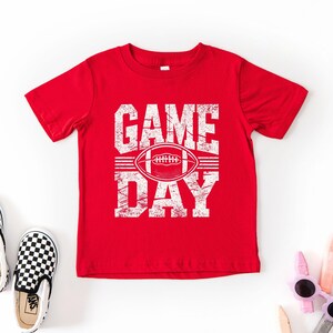 K&ouml;nnte beinhalten: Ein rotes T-Shirt mit den Worten "GAME DAY" in abgenutzten wei&szlig;en Buchstaben und einer Football-Grafik. Das Shirt wird mit schwarz-wei&szlig; karierten Slip-On-Schuhen und Sonnenbrillen kombiniert.