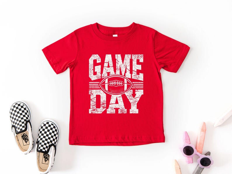 K&ouml;nnte beinhalten: Ein rotes T-Shirt mit den Worten "GAME DAY" in abgenutzten wei&szlig;en Buchstaben und einer Football-Grafik. Das Shirt wird mit schwarz-wei&szlig; karierten Slip-On-Schuhen und Sonnenbrillen kombiniert.
