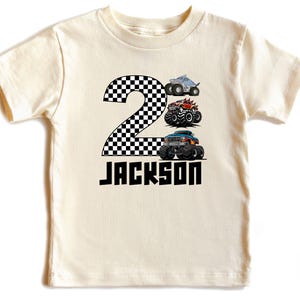 May include: Cream-colored toddler t-shirt with a black and white checkered number 2. Below the number are three monster truck illustrations and the name "JACKSON" in black block letters. A fun design for a child's birthday.