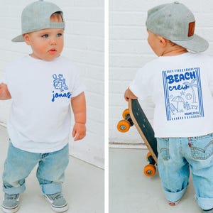 Op de afbeelding: Een wit t-shirt met blauwe graphics en de naam "jonas" in blauw schrift. Het shirt heeft een strandthema-ontwerp op de voorkant en de woorden "Beach Crew Miami 2024" op de achterkant. Het kind draagt een blauwe spijkerbroek en een grijze baseballcap.