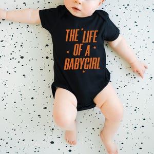 Pode incluir: Um body preto para beb&eacute; com o texto "THE LIFE OF A BABYGIRL" em laranja. O beb&eacute; est&aacute; deitado numa superf&iacute;cie branca com salpicos pretos.
