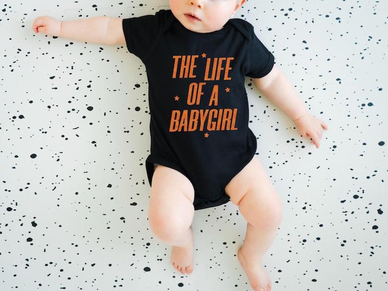 Pode incluir: Um body preto para beb&eacute; com o texto "THE LIFE OF A BABYGIRL" em laranja. O beb&eacute; est&aacute; deitado numa superf&iacute;cie branca com salpicos pretos.