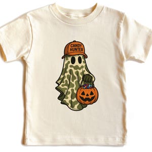 May include: Cream-colored t-shirt featuring a cartoon ghost wearing a brown cap with "CANDY HUNTER" text. The ghost is draped in a camouflage-patterned sheet and holds a jack-o'-lantern bucket filled with candy. A fun Halloween-themed design.