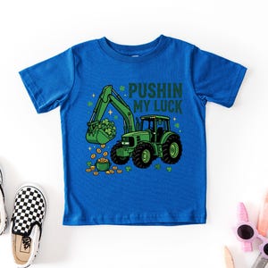 May include: A blue t-shirt featuring a green tractor with a bucket full of shamrocks and gold coins. The text "PUSHIN MY LUCK" is above the tractor. Black and white checkered slip-on shoes are next to the shirt.