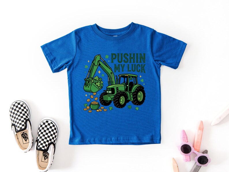 May include: A blue t-shirt featuring a green tractor with a bucket full of shamrocks and gold coins. The text "PUSHIN MY LUCK" is above the tractor. Black and white checkered slip-on shoes are next to the shirt.