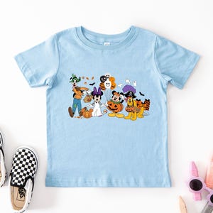 May include: Light blue t-shirt featuring a Halloween-themed graphic with Disney characters dressed in costumes. The graphic includes Mickey Mouse, Minnie Mouse, Goofy, Pluto, and other characters with pumpkins and bats. A pair of black and white checkered Vans shoes are next to the shirt.