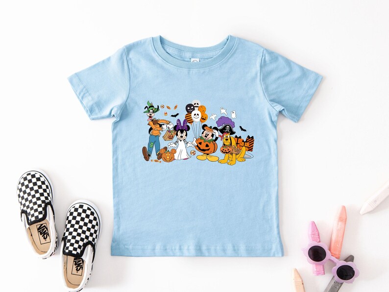 May include: Light blue t-shirt featuring a Halloween-themed graphic with Disney characters dressed in costumes. The graphic includes Mickey Mouse, Minnie Mouse, Goofy, Pluto, and other characters with pumpkins and bats. A pair of black and white checkered Vans shoes are next to the shirt.