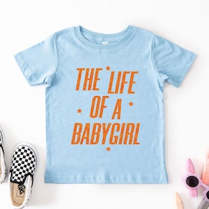 Pode incluir: Camiseta azul claro com o texto laranja "THE LIFE OF A BABYGIRL". A imagem tamb&eacute;m mostra um par de sapatos slip-on xadrez em preto e branco. &Oacute;culos de sol e l&aacute;pis de cor tamb&eacute;m s&atilde;o vis&iacute;veis.