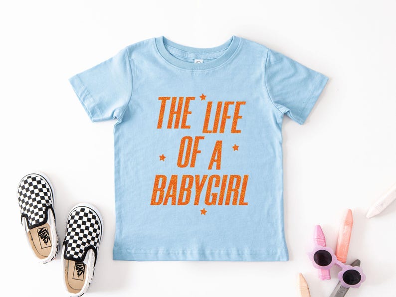 Pode incluir: Camiseta azul claro com o texto laranja "THE LIFE OF A BABYGIRL". A imagem tamb&eacute;m mostra um par de sapatos slip-on xadrez em preto e branco. &Oacute;culos de sol e l&aacute;pis de cor tamb&eacute;m s&atilde;o vis&iacute;veis.