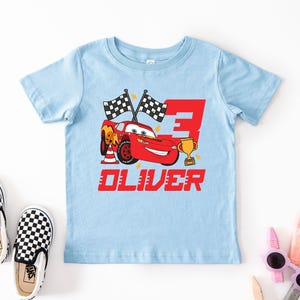 May include: A light blue toddler t-shirt featuring a red cartoon race car with the number 3, checkered flags, and a gold trophy. The name "OLIVER" is printed in red below the car. Black and white checkered slip-on shoes are next to the shirt.