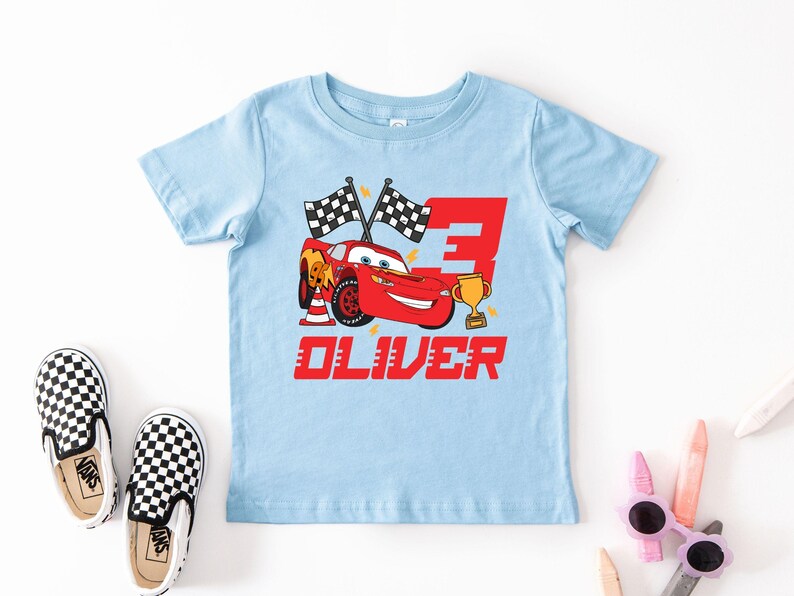 May include: A light blue toddler t-shirt featuring a red cartoon race car with the number 3, checkered flags, and a gold trophy. The name "OLIVER" is printed in red below the car. Black and white checkered slip-on shoes are next to the shirt.